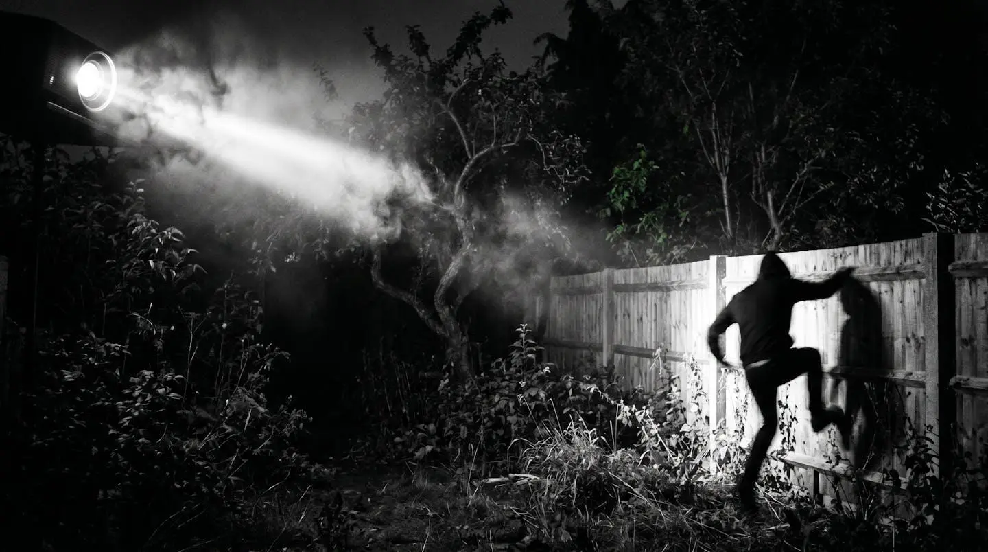 Jardin de nuit éclairé par un projecteur, une silhouette floue s'enfuit.